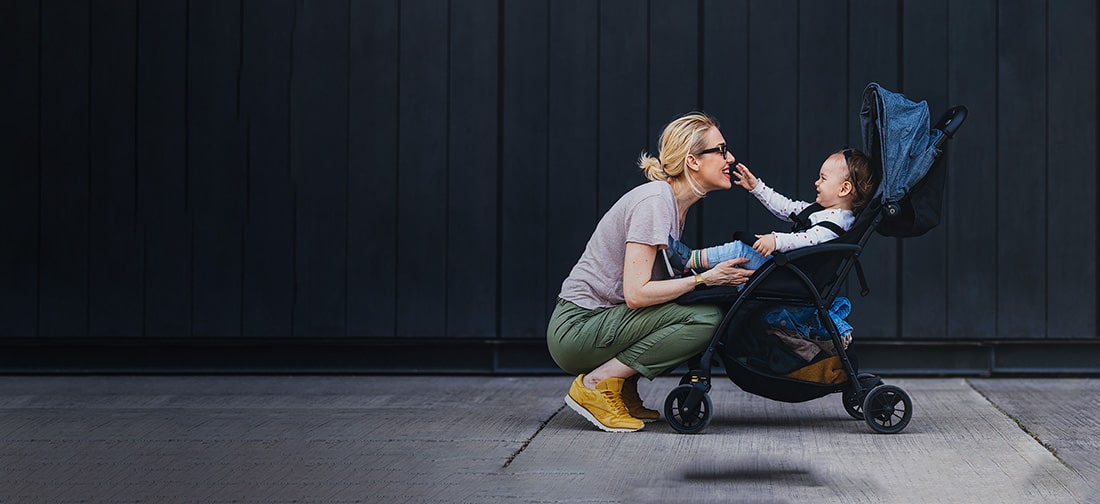 Mother squatting in front of her child in a stroller as the child reaches out for her. 