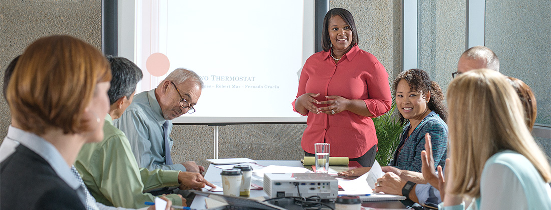 Multi-generational employees seated around desk listen to presentation.