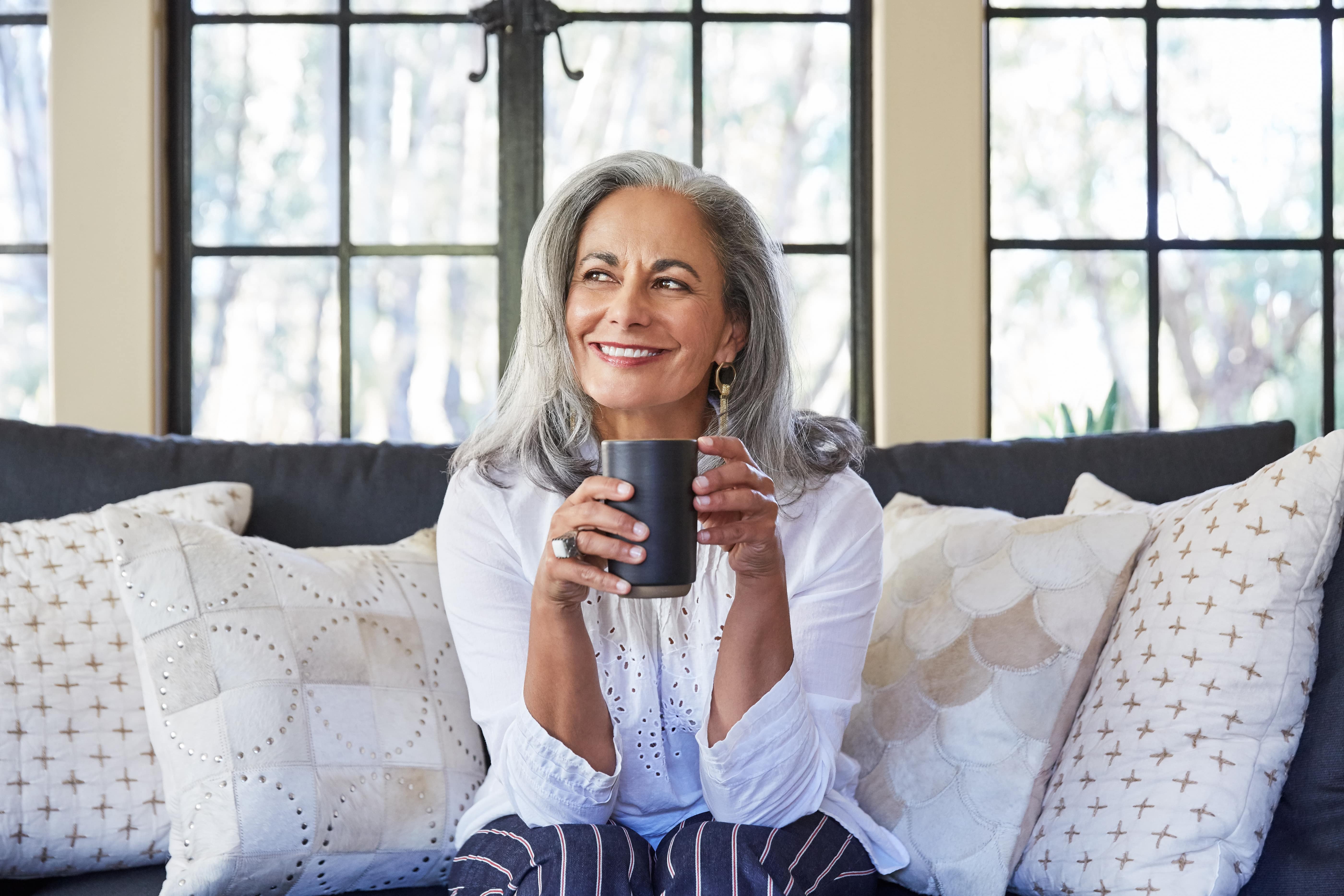 Older woman sitting in the couch, smiling and holding a cup of coffee